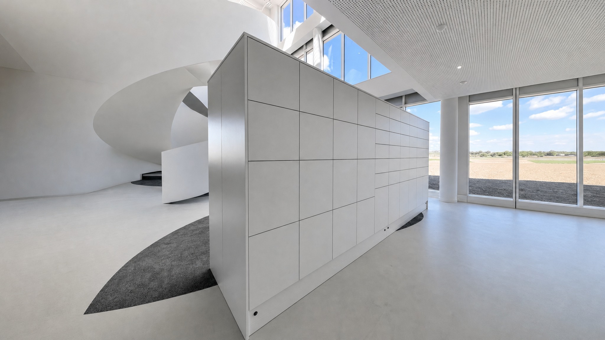 Large bank of light gray lockers in spacious modern room with curved stairs and large windows.