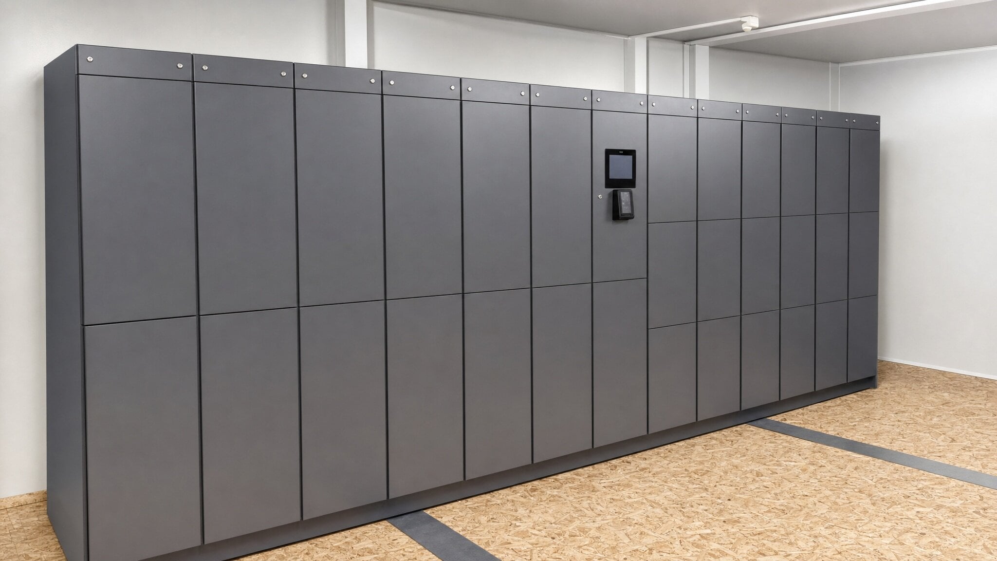 Gray locker bank with integrated display and access panel in a plain indoor room.