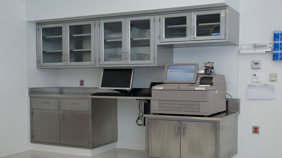 Durable metal casework system featuring glass-door wall cabinets and integrated computer workstation in a clinical or pharmacy setting.