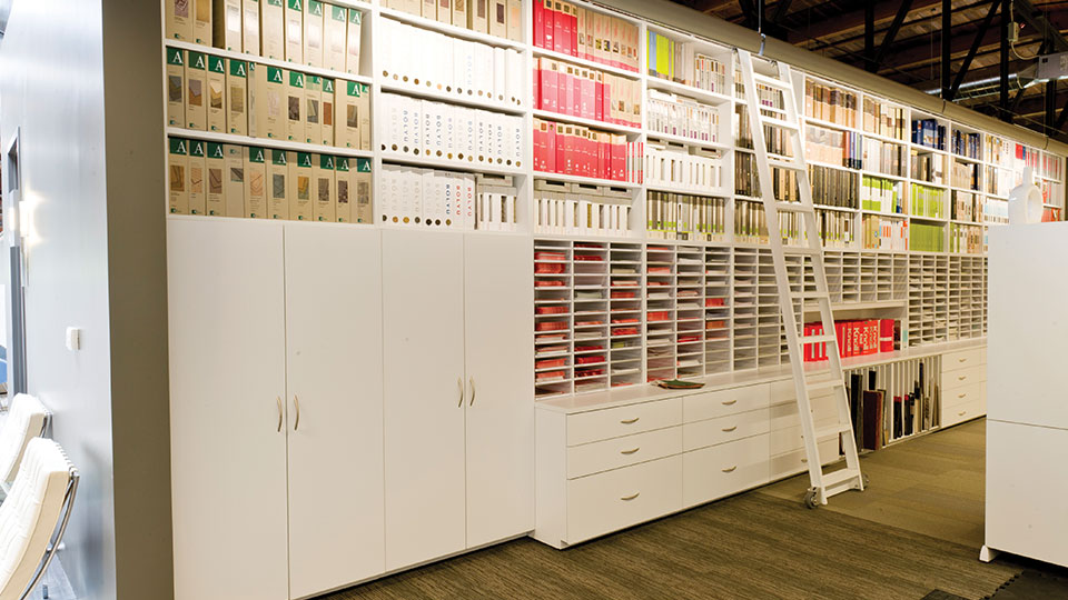 High-density modular millwork display wall with vertical shelving, sample bins, and a rolling ladder for design libraries and architectural firms.